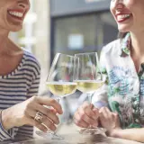 Två glada kvinnor skålar med vitt vin. En titt på kalorier alkohol tabell kan vara bra att ha i åtanke.
