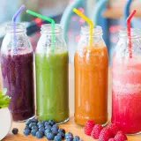 Fyra flaskor med nyttig smoothie i olika färger: lila, grön, orange och rosa. Blåbär och hallon ligger bredvid.