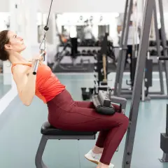 Kvinna utför enarms latsdrag på gym. Hon sitter på en bänk och drar ner en stång med ett rep.