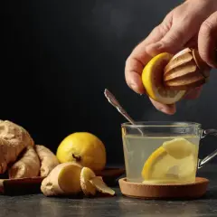 En hand pressar citron i en kopp med varmt citronvatten på morgonen. Ingefära och citroner ligger bredvid.