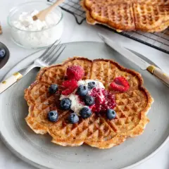 En gyllene protein våffla toppad med bär och grädde. Serveras med en gaffel och kniv, redo att avnjutas.