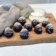 Mörka, chokladöverdragna protein chokladbollar toppade med kokosflingor ligger på en träskärbräda.
