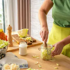Person in green shirt and yellow apron packing shredded cabbage into a glass jar with a wooden tamper.