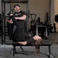 En kvinna utför sidoliggande benlyft på en bänk i ett gym, med en tränare som observerar.