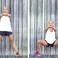 Kvinna utför sumo squat framför trävägg. Bredbent, djup knäböj med armarna framåt.