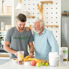 Två män i köket förbereder en smoothie med frukt. En burk med proteinpulver står bredvid, perfekt för att täcka ditt proteinbehov per dag.