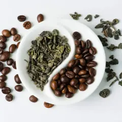 En vit skål delad i två, med torkade gröna teblad på ena sidan och rostade kaffebönor på den andra. En visuell jämförelse av koffein i te vs kaffe.