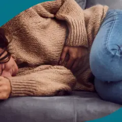 En kvinna med mörkt hår ligger hopkrupen på en soffa, med handen på magen. Hon ser ut att ha ont, vilket kan vara ett av många endometrios symtom.