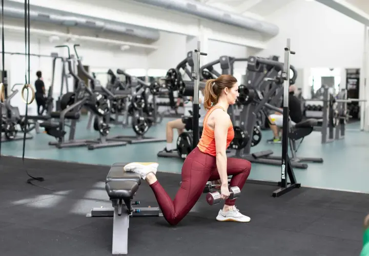 Kvinna utför bulgarian split squat med hantlar, tränar benmuskler och rumpa i gym.