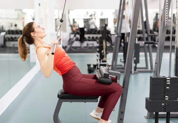 Kvinna utför enarms latsdrag på gym. Hon sitter på en bänk och drar ner en stång med ett rep.