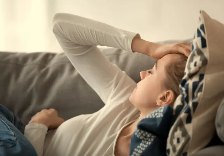 Kvinna med handen på pannan, kanske ett av b12-brist symtom som trötthet eller huvudvärk. Hon vilar på en soffa.