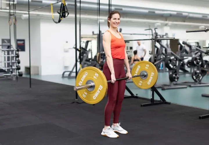 Kvinna utför marklyft med skivstång i gym. Hon har en orange topp och vinröda träningstights.