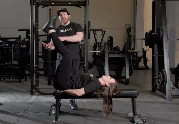 En kvinna utför sidoliggande benlyft på en bänk i ett gym, med en tränare som observerar.