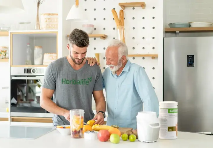 Två män i köket förbereder en smoothie med frukt. En burk med proteinpulver står bredvid, perfekt för att täcka ditt proteinbehov per dag.