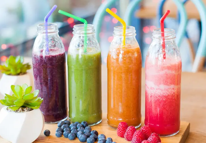 Fyra flaskor med nyttig smoothie i olika färger: lila, grön, orange och rosa. Blåbär och hallon ligger bredvid.