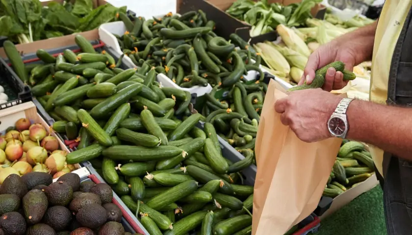 En person lägger gurka nyttigt i en papperspåse på en marknad. Gurkorna ligger i en stor låda, bredvid päron och avokado.