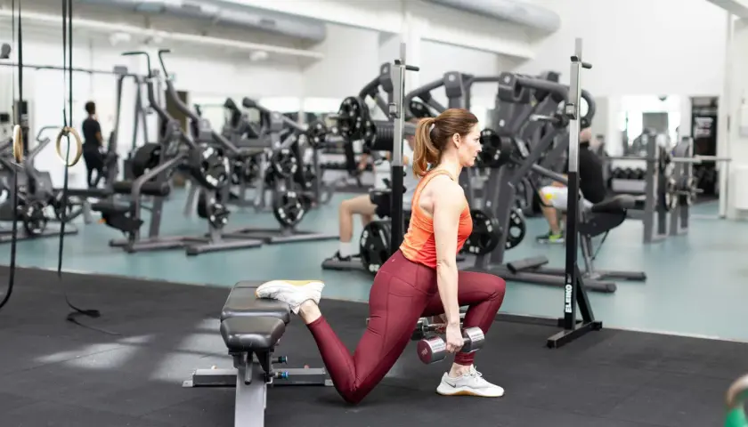 Kvinna utför bulgarian split squat med hantlar, tränar benmuskler och rumpa i gym.