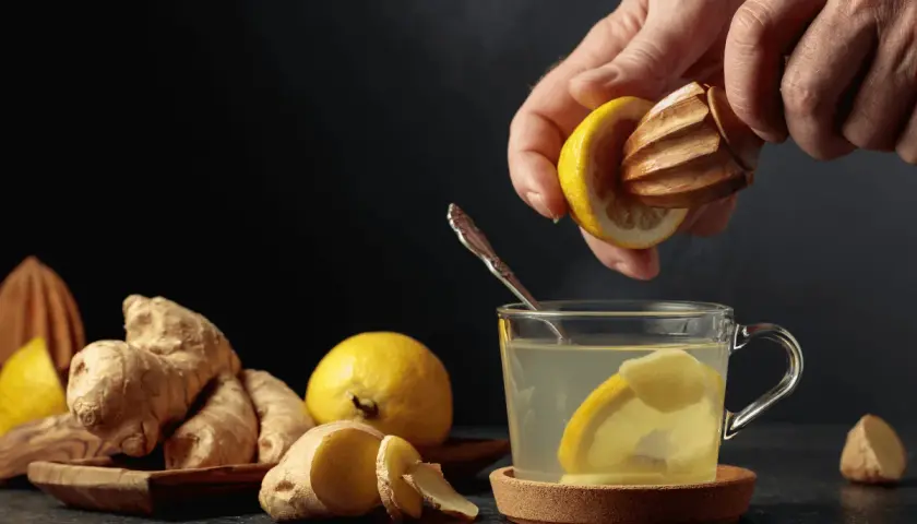 En hand pressar citron i en kopp med varmt citronvatten på morgonen. Ingefära och citroner ligger bredvid.