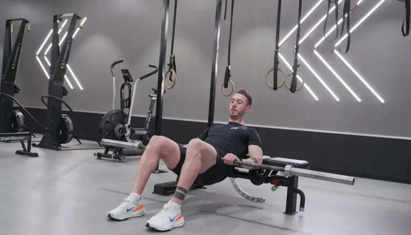 Man utför höftlyft med skivstång för att bygga muskler i rumpan. Träningspasset sker i ett modernt gym.