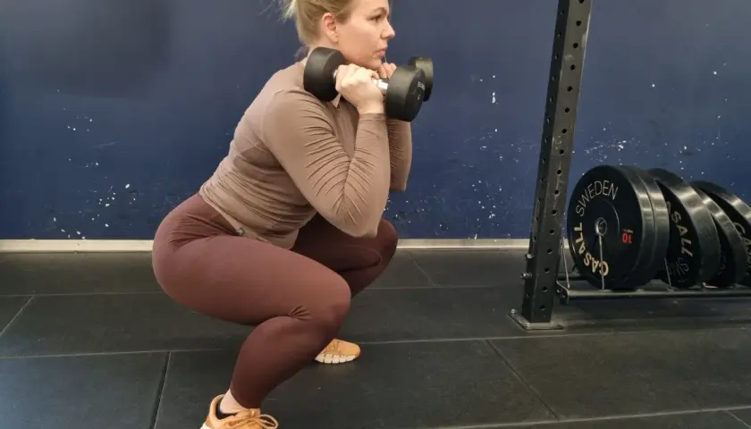 Kvinna utför squats med hantlar i ett gym. Hon har en brun tröja och bruna tights.