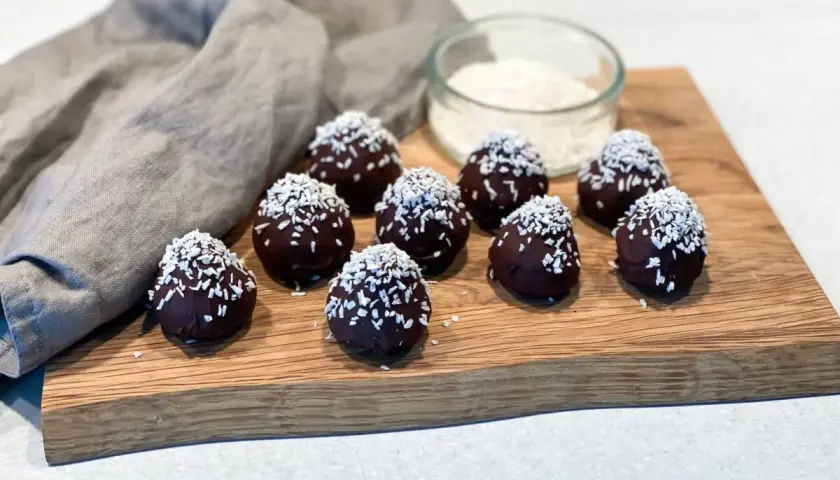 Mörka, chokladöverdragna protein chokladbollar toppade med kokosflingor ligger på en träskärbräda.