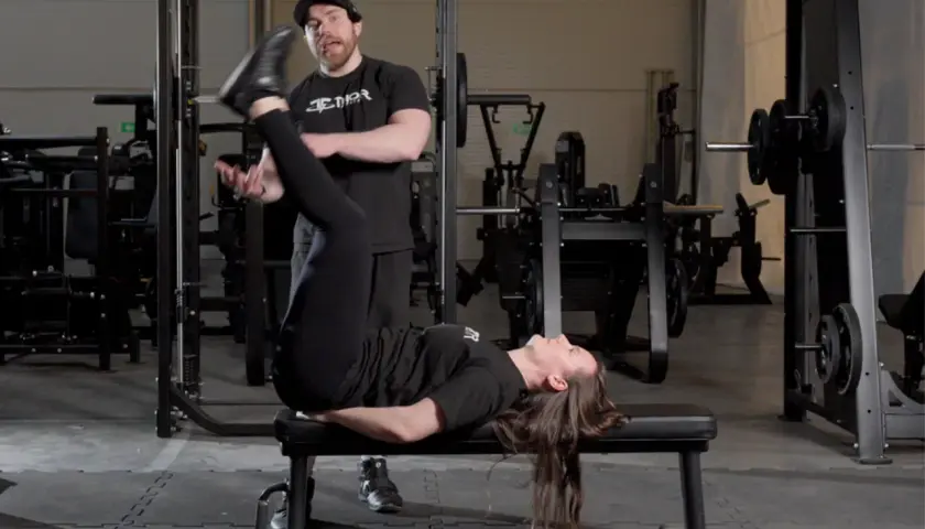 En kvinna utför sidoliggande benlyft på en bänk i ett gym, med en tränare som observerar.