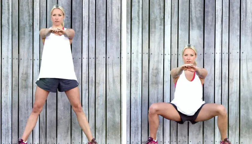 Kvinna utför sumo squat framför trävägg. Bredbent, djup knäböj med armarna framåt.