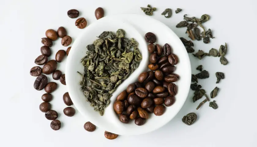 En vit skål delad i två, med torkade gröna teblad på ena sidan och rostade kaffebönor på den andra. En visuell jämförelse av koffein i te vs kaffe.