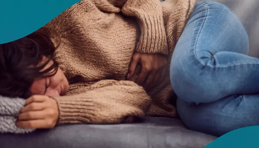 En kvinna med mörkt hår ligger hopkrupen på en soffa, med handen på magen. Hon ser ut att ha ont, vilket kan vara ett av många endometrios symtom.