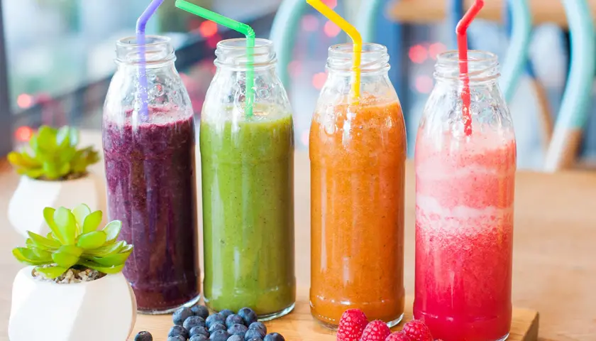 Fyra flaskor med nyttig smoothie i olika färger: lila, grön, orange och rosa. Blåbär och hallon ligger bredvid.