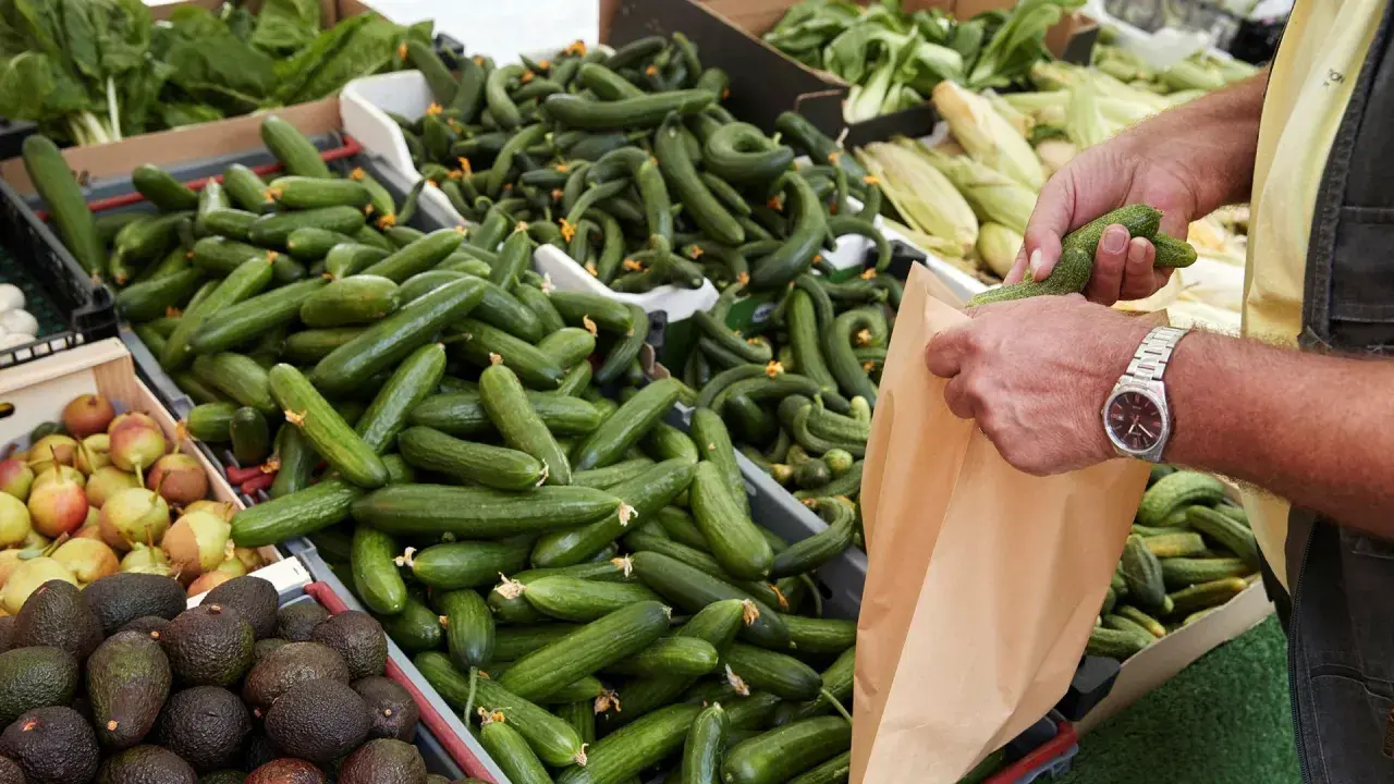 En person lägger gurka nyttigt i en papperspåse på en marknad. Gurkorna ligger i en stor låda, bredvid päron och avokado.