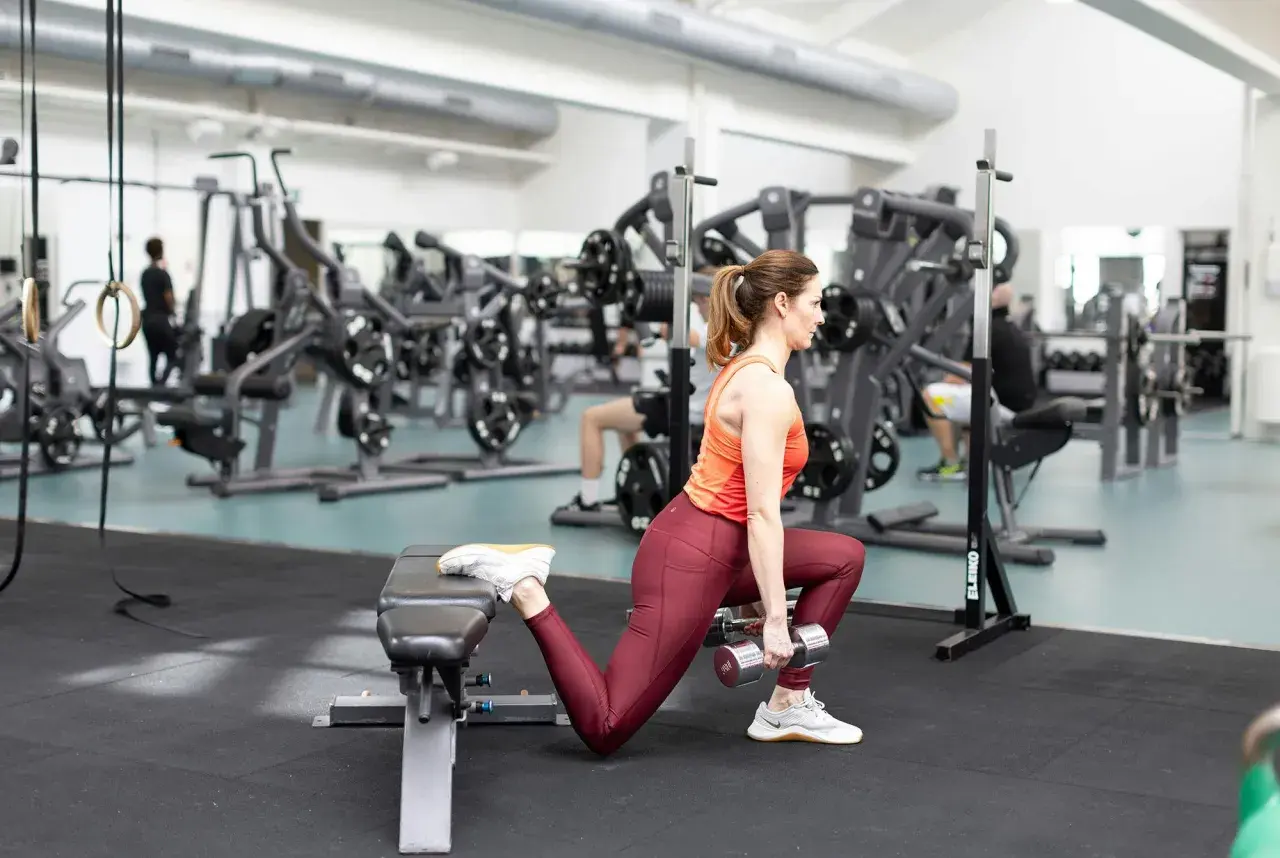 Kvinna utför bulgarian split squat med hantlar, tränar benmuskler och rumpa i gym.
