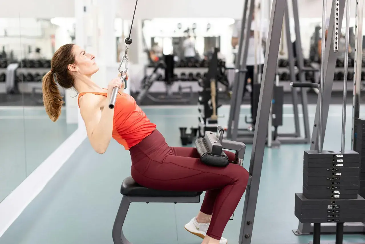 Kvinna utför enarms latsdrag på gym. Hon sitter på en bänk och drar ner en stång med ett rep.