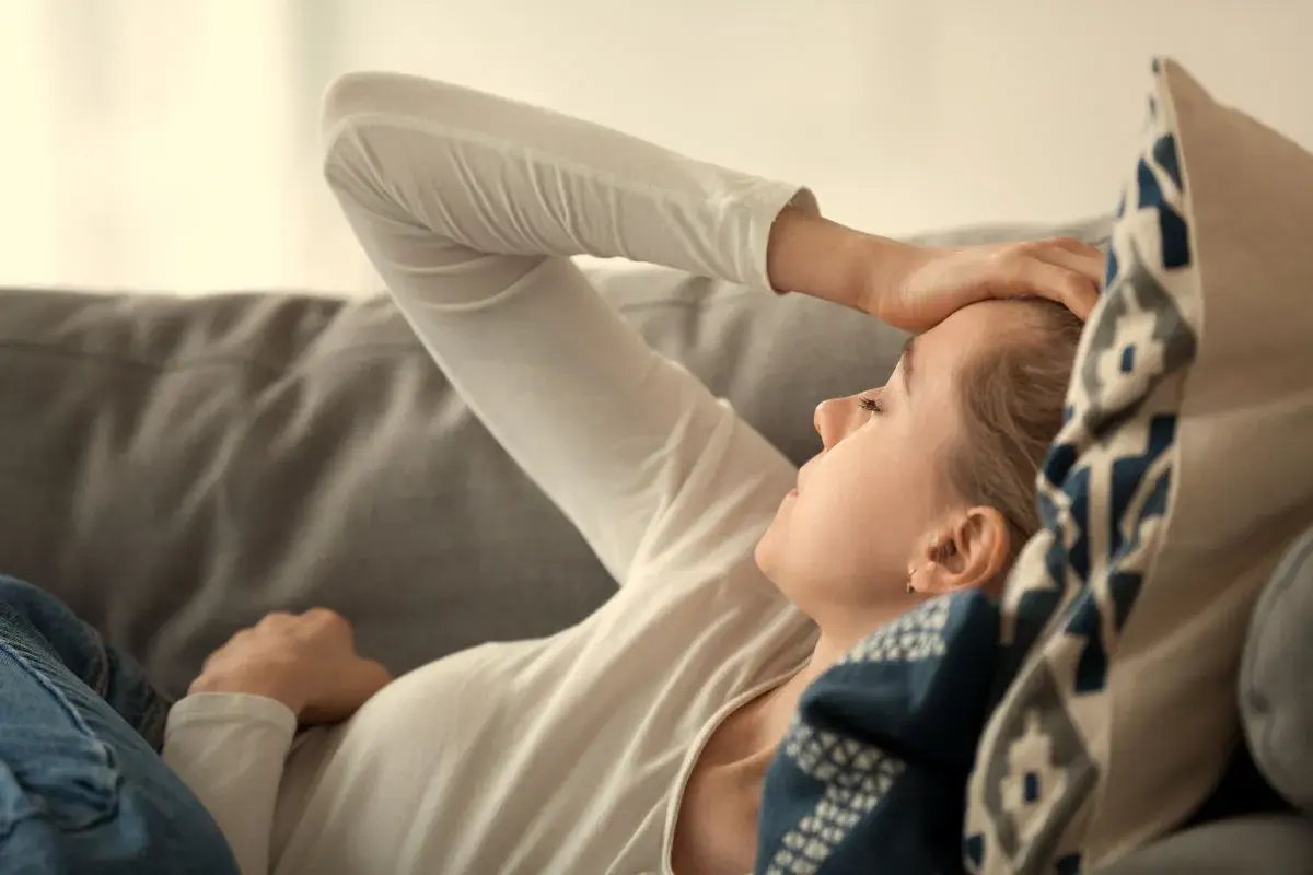 Kvinna med handen på pannan, kanske ett av b12-brist symtom som trötthet eller huvudvärk. Hon vilar på en soffa.