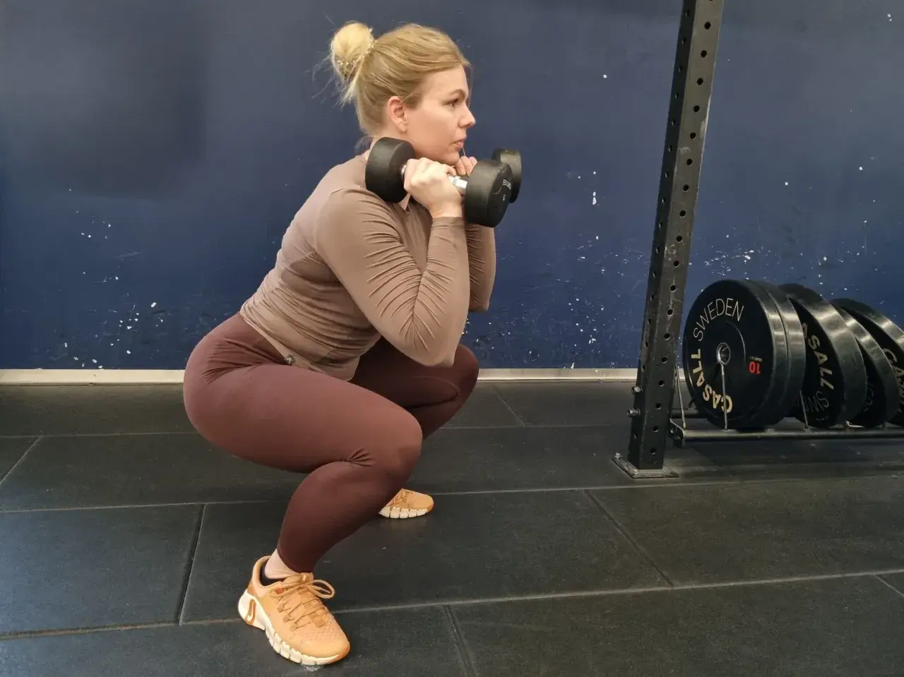 Kvinna utför squats med hantlar i ett gym. Hon har en brun tröja och bruna tights.