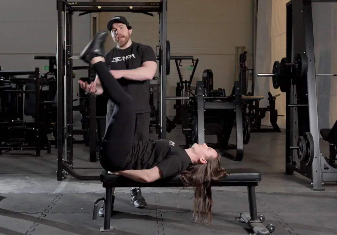 En kvinna utför sidoliggande benlyft på en bänk i ett gym, med en tränare som observerar.