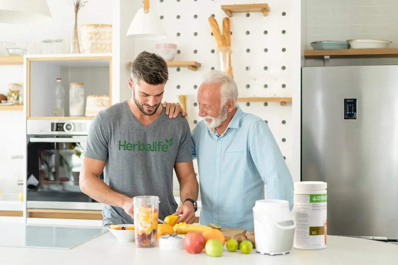 Två män i köket förbereder en smoothie med frukt. En burk med proteinpulver står bredvid, perfekt för att täcka ditt proteinbehov per dag.