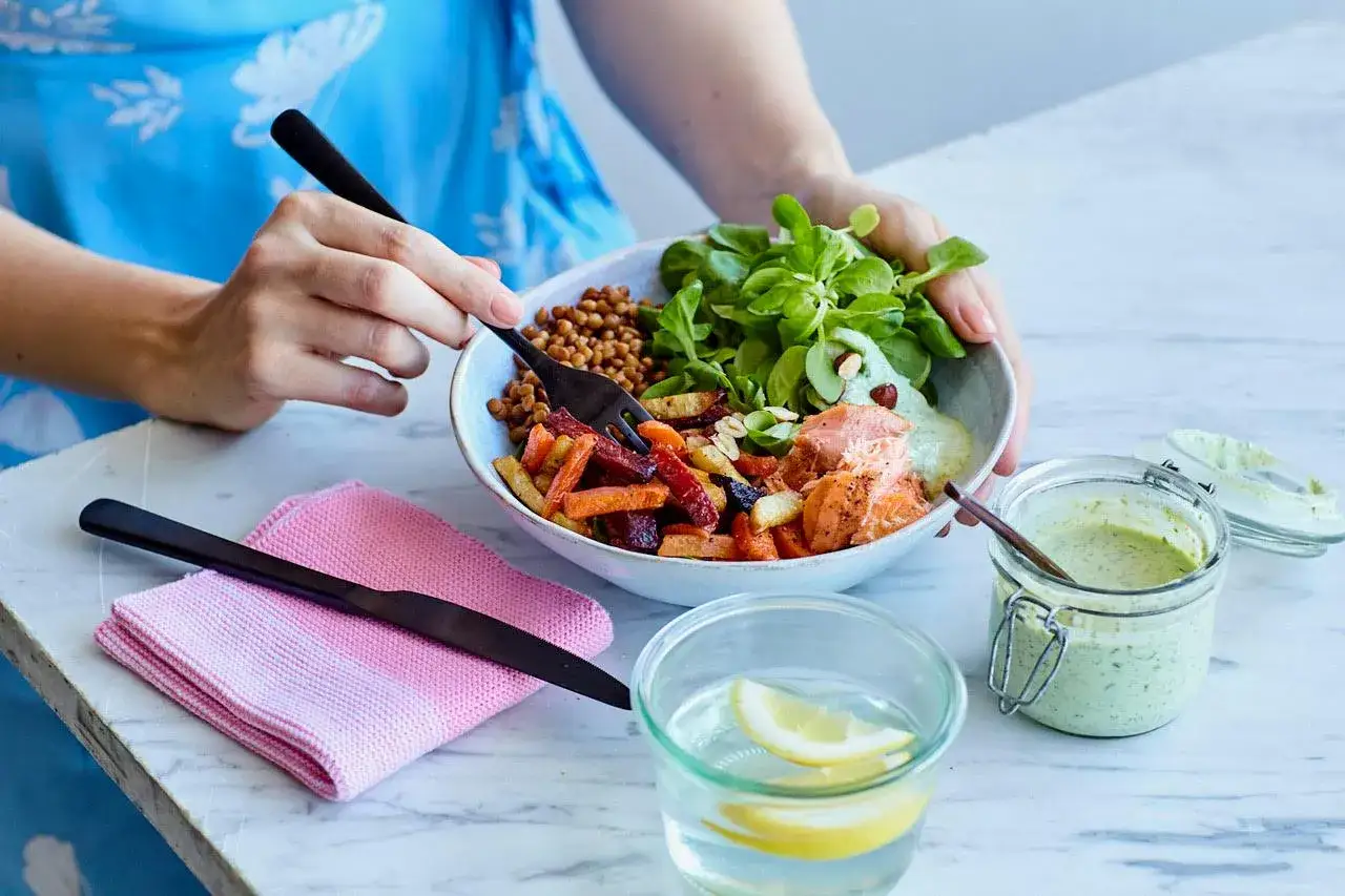 En person njuter av en nyttig middag med lax, rotfrukter, linser och gr&ouml;nsallad. En gr&ouml;n dressing st&aring;r bredvid.