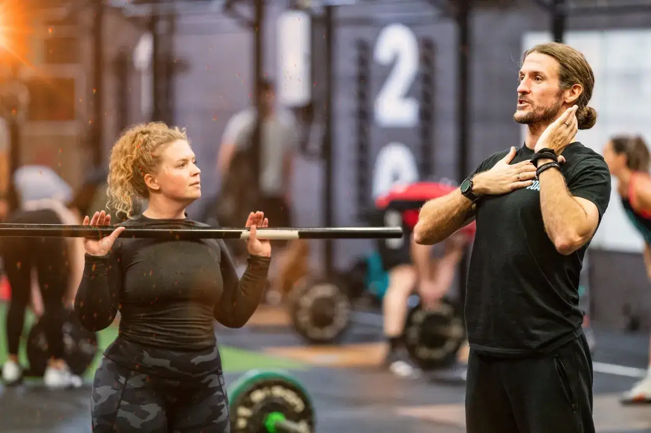 En kvinna f&ouml;rbereder sig f&ouml;r ett lyft med en skivst&aring;ng under ett crossfit pass. Tr&auml;naren ger instruktioner.