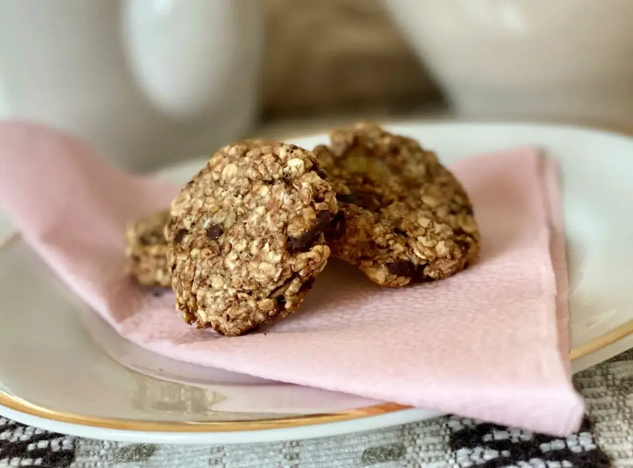 Tre hembakade nyttiga kakor med chokladbitar ligger på en ljusrosa servett på en vit tallrik med guldkant.
