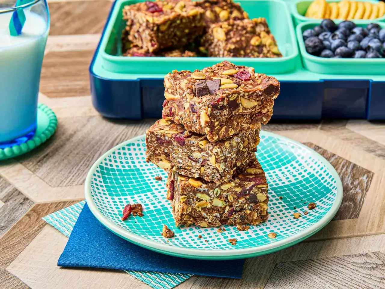 En stapel hemmagjorda proteinbars med nötter, torkad frukt och chokladbitar. En matlåda med blåbär och kex syns i bakgrunden.