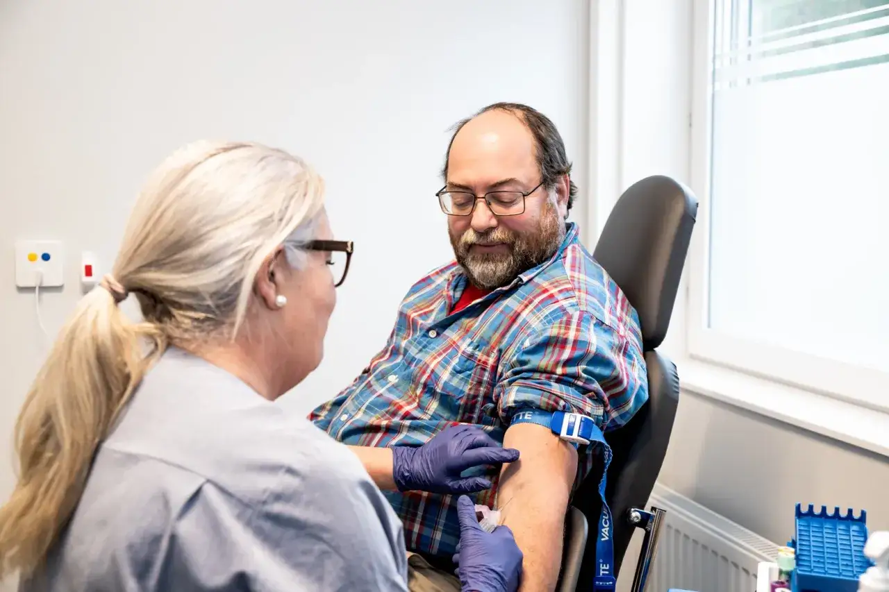Sjuksköterska tar blodprov på man som sitter i stol. Mannen har glasögon och rutig skjorta. Han har fasta innan blodprov.