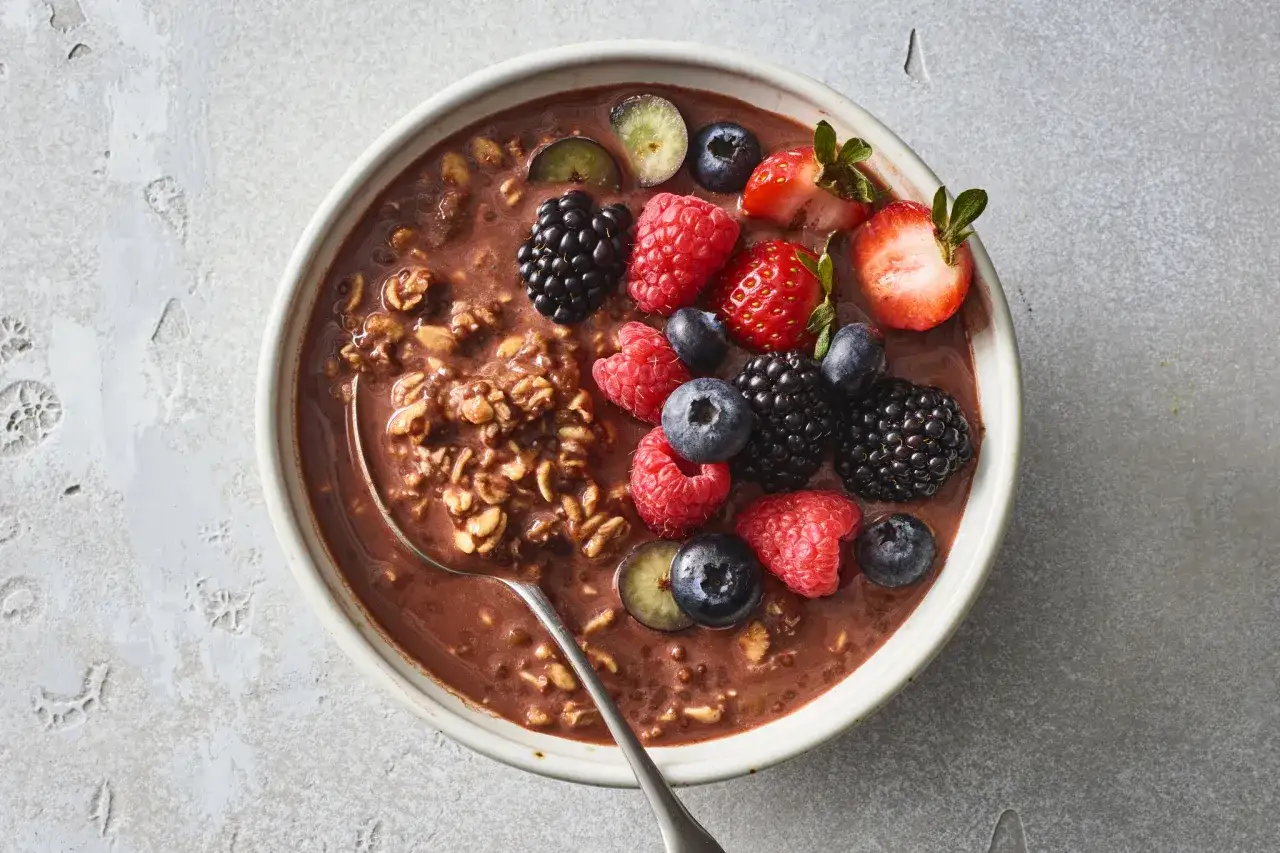 En sk&aring;l med overnight oats kakao, toppad med f&auml;rska hallon, bl&aring;b&auml;r, bj&ouml;rnb&auml;r och jordgubbar. En sked ligger i sk&aring;len.